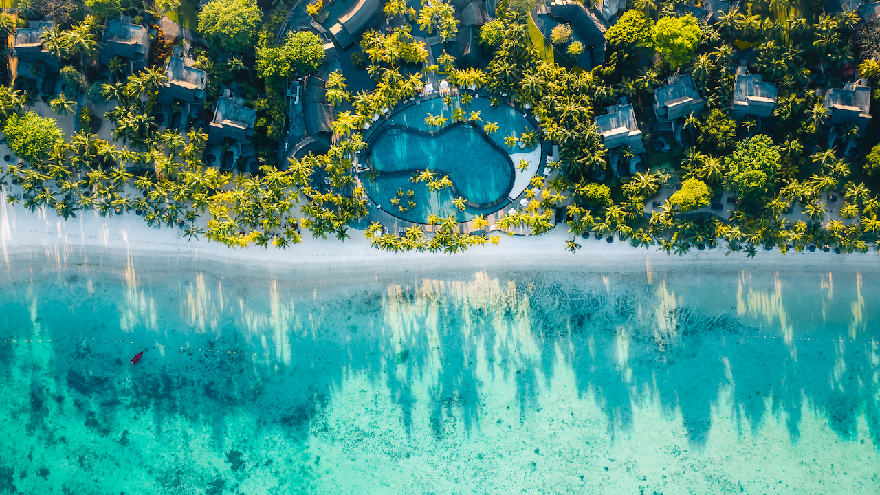 Vista aerea di un resort tropicale sulla spiaggia con una grande piscina circondata da palme, spiaggia di sabbia bianca, acqua turchese dell'oceano ed edifici sparsi immersi in una vegetazione lussureggiante.