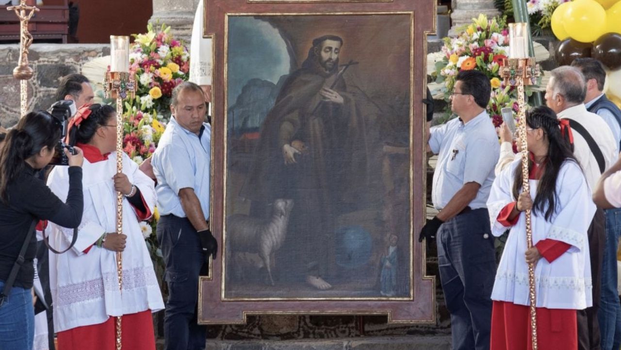 Diverse persone, tra cui i servitori dell'altare in abiti rossi e bianchi, stanno intorno a un grande quadro religioso incorniciato che raffigura un santo con un agnello e lo tengono in piedi all'aperto, circondato da composizioni floreali.