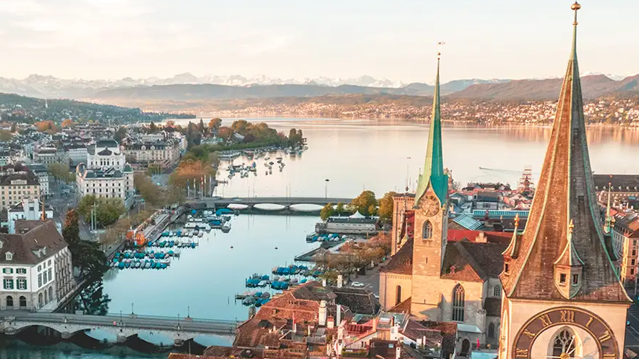 Una vista panoramica di Zurigo, in Svizzera, con le acque serene del lago, le barche, gli edifici storici e le guglie delle chiese sullo sfondo delle montagne, che evocano il fascino di Lugano sotto un cielo limpido.