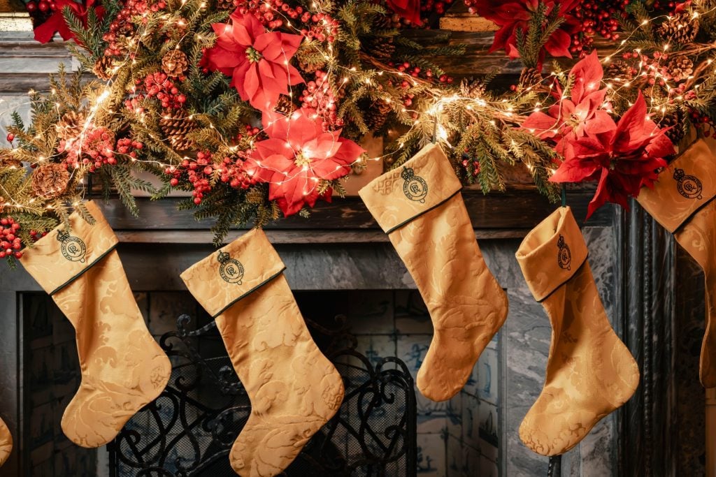 Cinque calze di Natale dorate pendono da una ghirlanda decorata sul camino con stelle di Natale rosse, bacche, rami di pino e luci scintillanti.