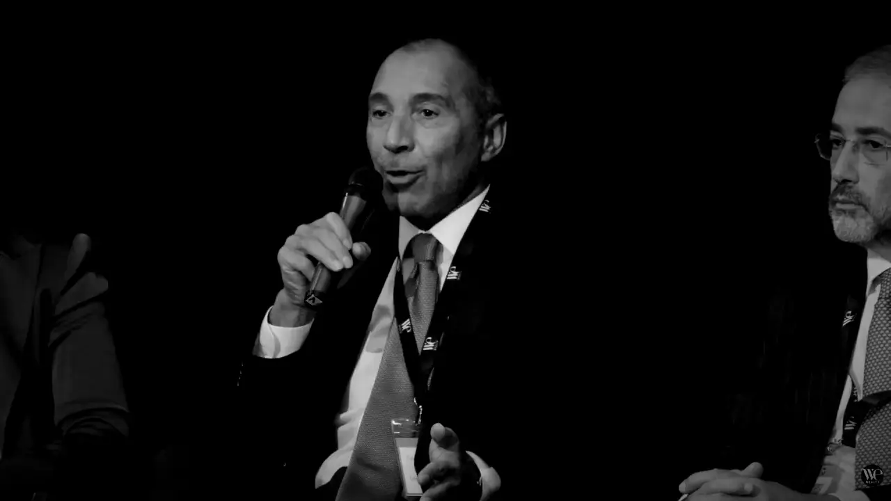 Un uomo in giacca e cravatta parla al microfono durante un evento, seduto tra altre persone parzialmente visibili su uno sfondo scuro.