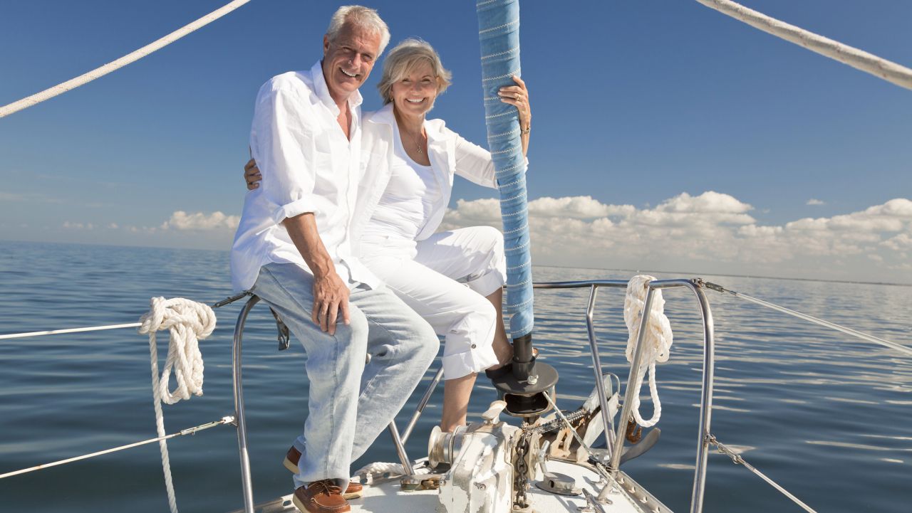 Una coppia di anziani in abiti bianchi siede e sorride insieme a prua di una barca a vela su acque calme, con un cielo blu e nuvole sparse sullo sfondo.