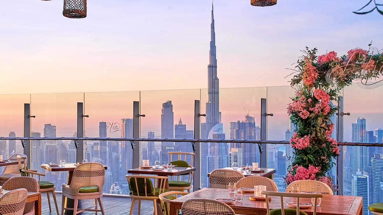 A rooftop restaurant with wicker chairs and wooden tables overlooks a city skyline at sunset, featuring the Burj Khalifa in the center and a floral arch on the right.