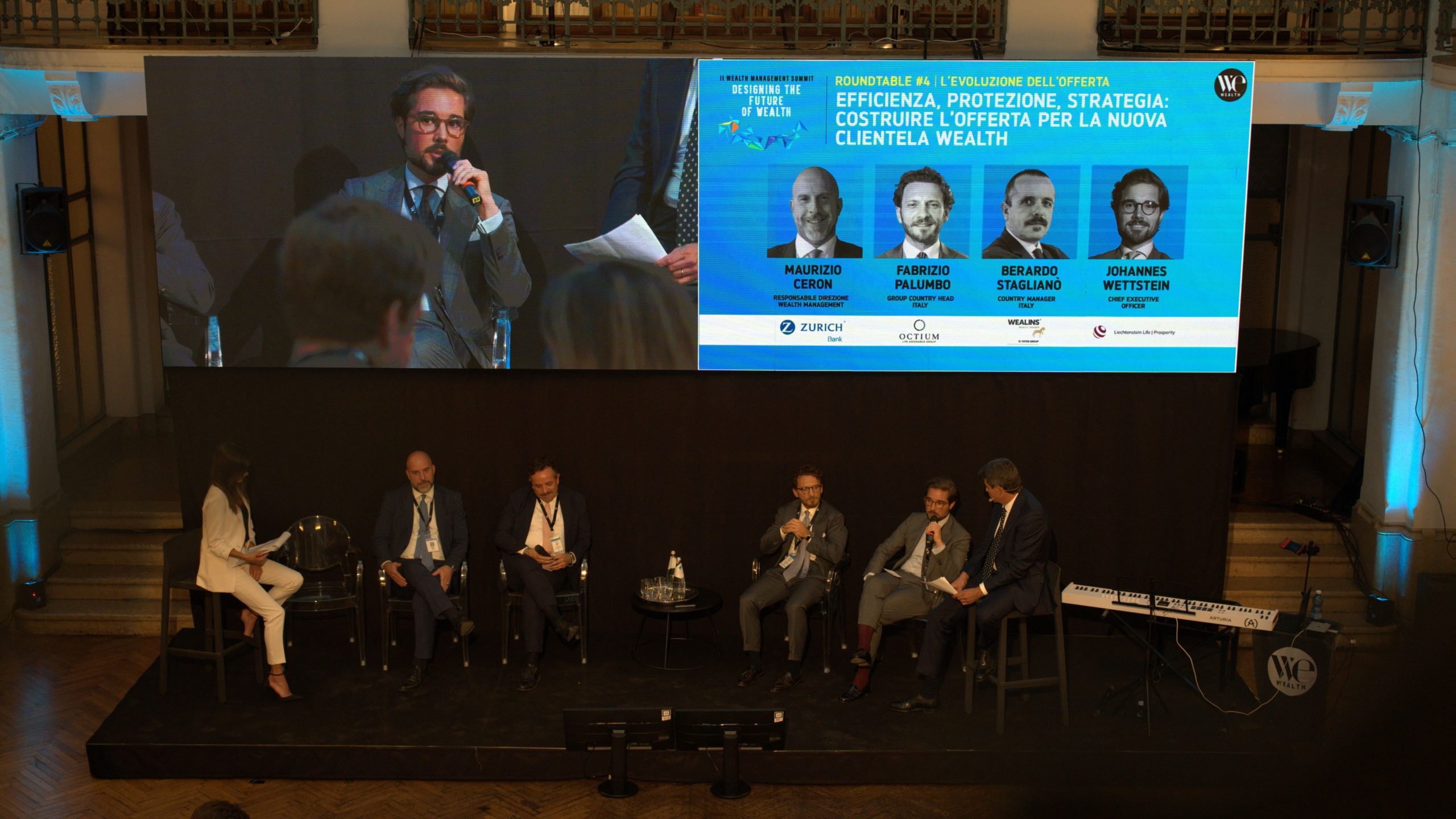 A panel of six people, five men in suits and one woman in white, sits on a stage discussing, with a large screen behind them displaying their names and event details in Italian. The setting is a formal conference.