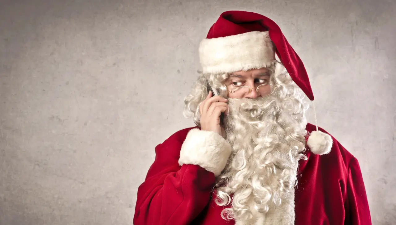 Babbo Natale con barba bianca e vestito rosso parla al cellulare, guardando di lato con espressione curiosa, su uno sfondo grigio.