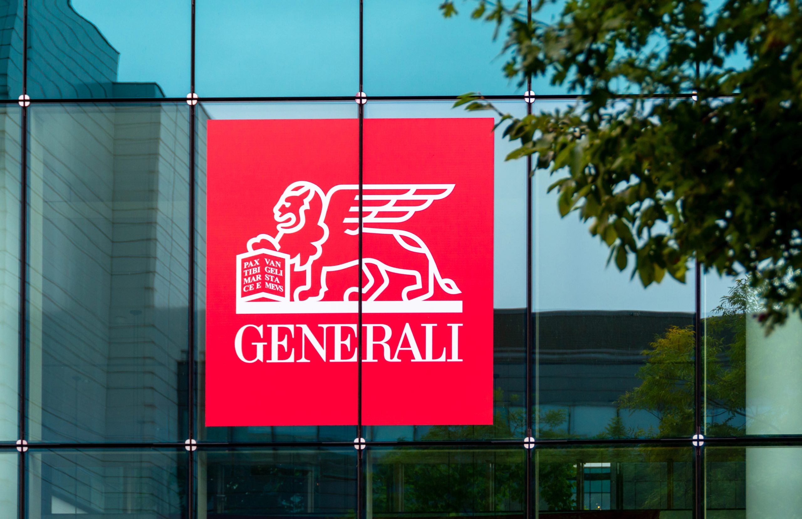 Una grande insegna rossa con il logo delle Generali, raffigurante un leone alato che regge un libro, campeggia sulla facciata in vetro di un edificio. Sullo sfondo sono visibili alberi e riflessi di altri edifici, tra cui Natixis.