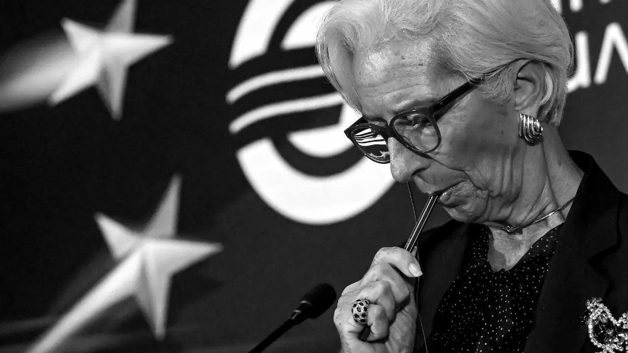Una donna anziana con gli occhiali guarda pensierosa, portandosi una penna alle labbra. È in piedi davanti a uno sfondo con stelle e un grande simbolo della valuta euro. L'immagine è in bianco e nero.