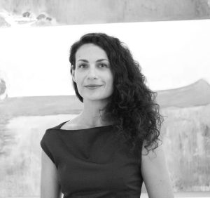 Una donna con i capelli ricci e scuri, che indossa un abito scuro, si trova di fronte a un grande dipinto astratto. L'immagine è in bianco e nero e la donna guarda la telecamera con un leggero sorriso.