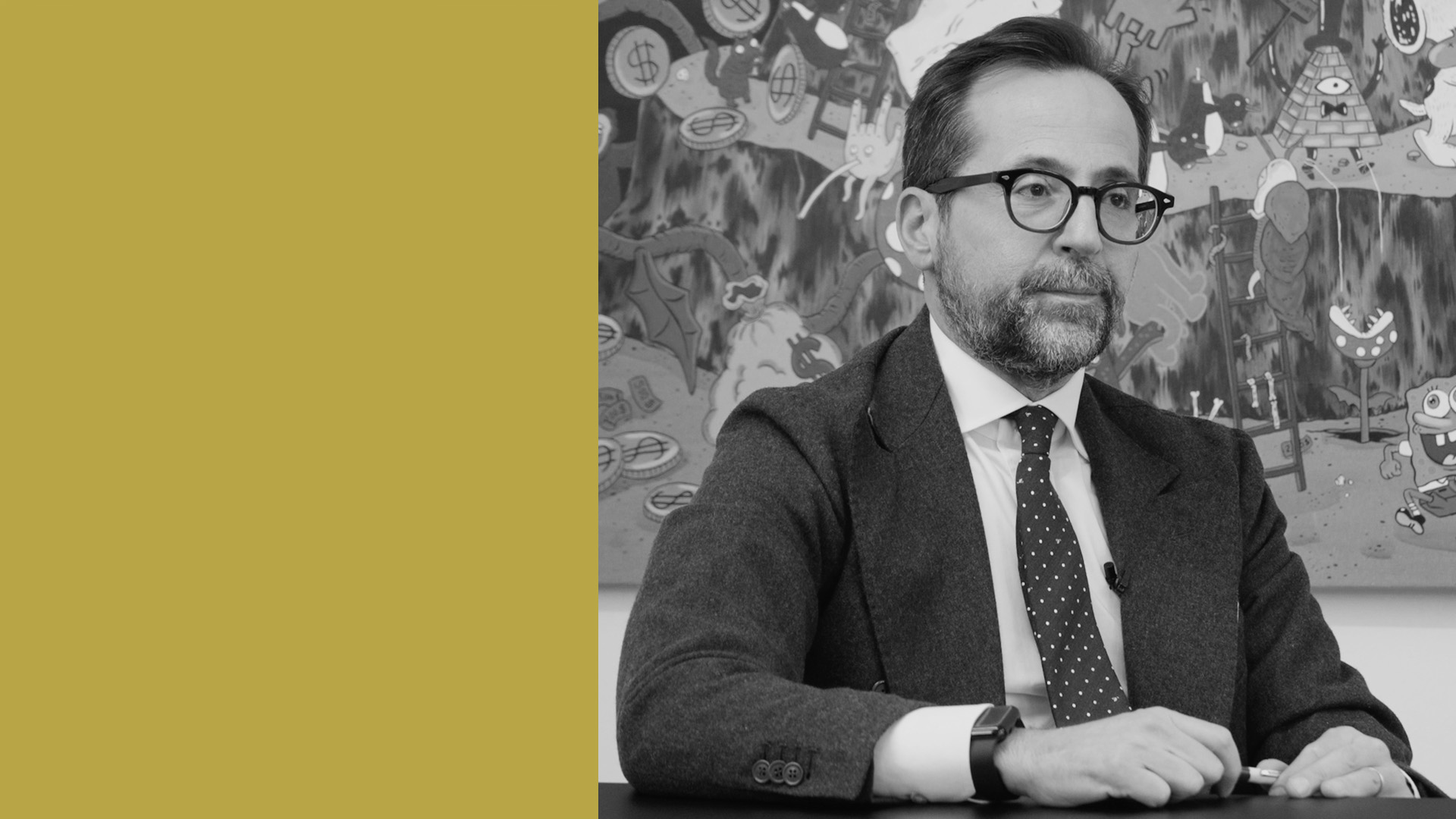 Un uomo con occhiali e barba indossa un abito e una cravatta a pois, seduto a un tavolo. Dietro di lui c'è un dipinto astratto colorato. Il lato sinistro dell'immagine è caratterizzato da un semplice sfondo dorato.