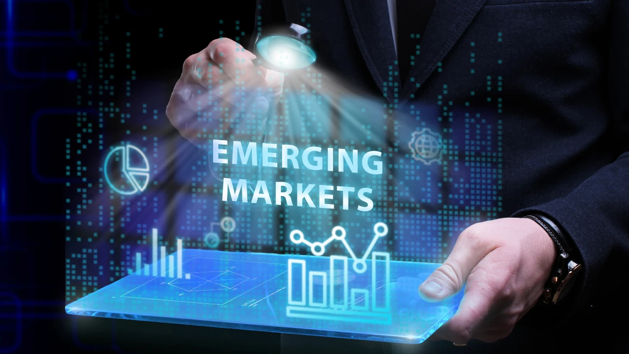 Una persona in giacca e cravatta tiene in mano un tablet che visualizza grafici e dati digitali. Le parole "EMERGING MARKETS" e "debito emergente" sono illuminate in alto, con icone futuristiche ed elementi luminosi blu sullo sfondo.