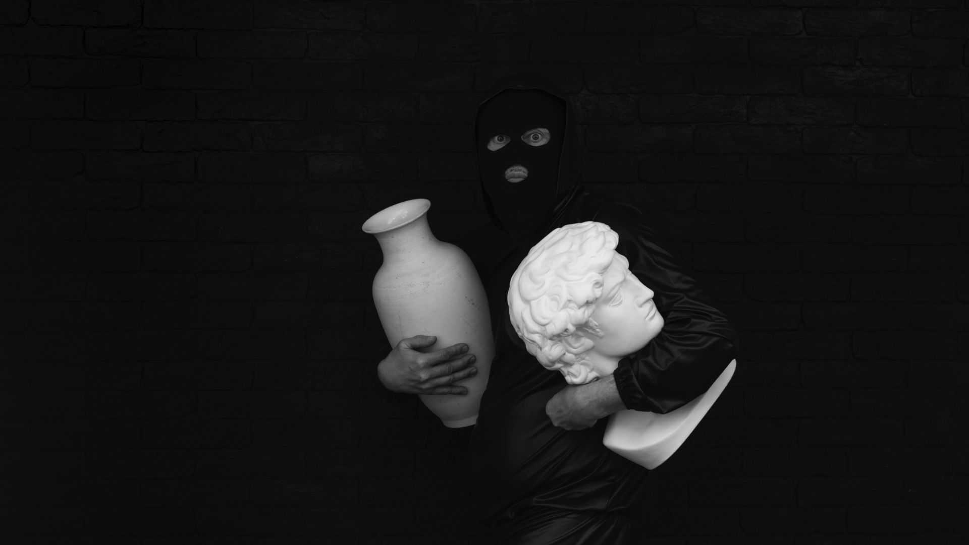 Una persona con passamontagna nero e abiti scuri, che evocano scene di furti d'arte, tiene in mano un vaso di ceramica bianca e un busto bianco in stile classico contro una parete di mattoni scuri.
