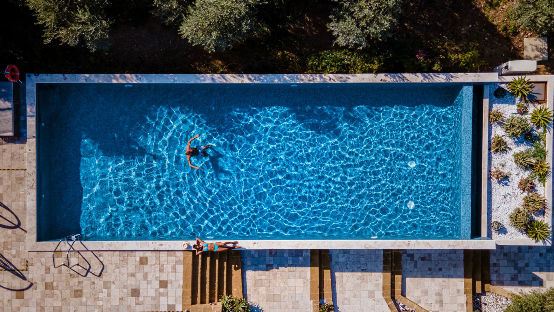 Vista aerea di una piscina rettangolare con una persona che nuota all'interno e un'altra seduta a bordo vasca; l'area circostante è caratterizzata da piastrelle in pietra e piante paesaggistiche.