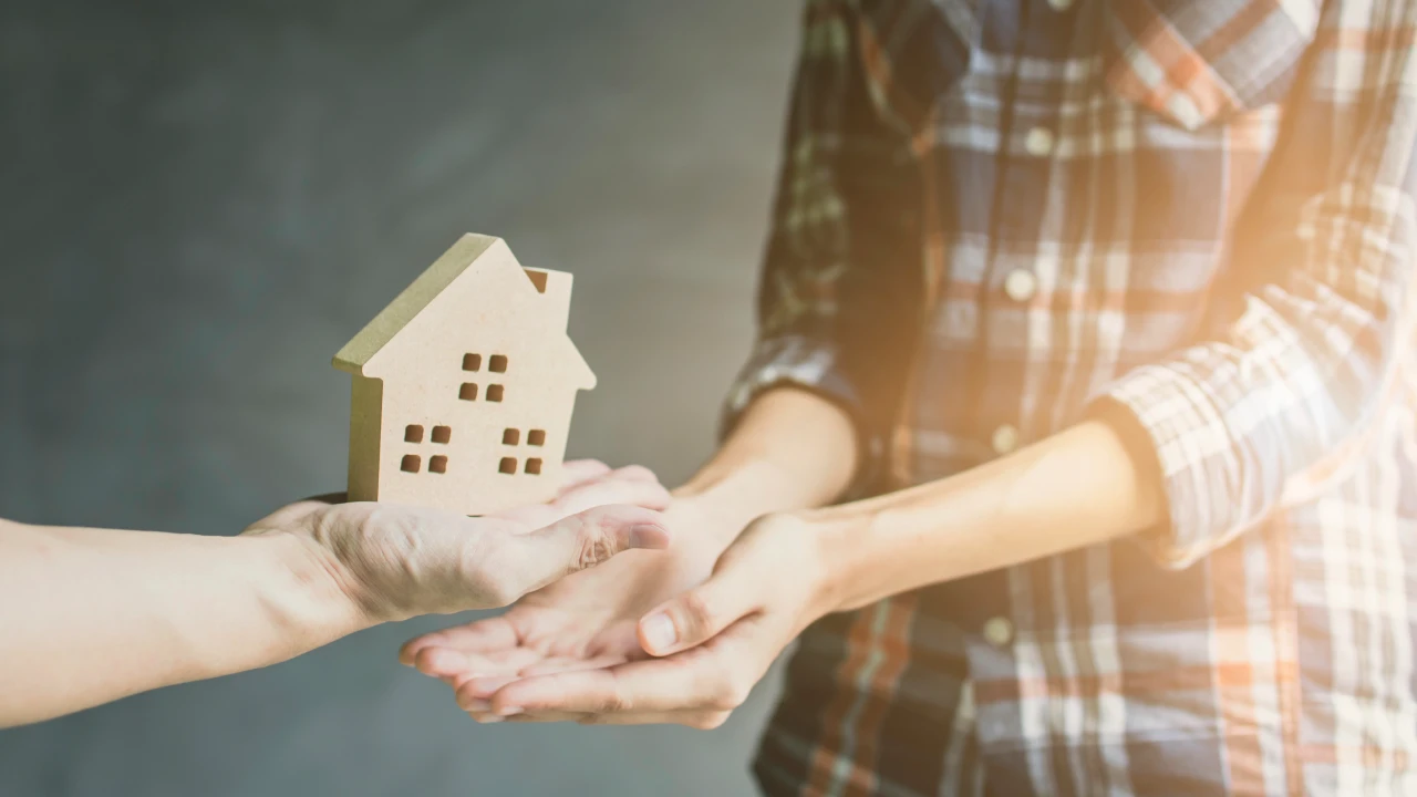 Due persone si scambiano un modellino di casa in legno; una mano lo offre e l'altra lo riceve. Lo sfondo è leggermente sfocato. Il riferimento va agli immobili donati.