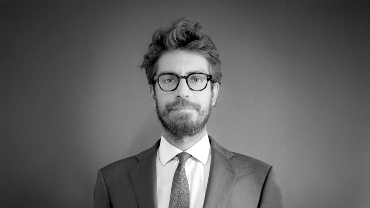 Un uomo con occhiali e barba, in giacca e cravatta, è in piedi su uno sfondo semplice. L'immagine è in bianco e nero.