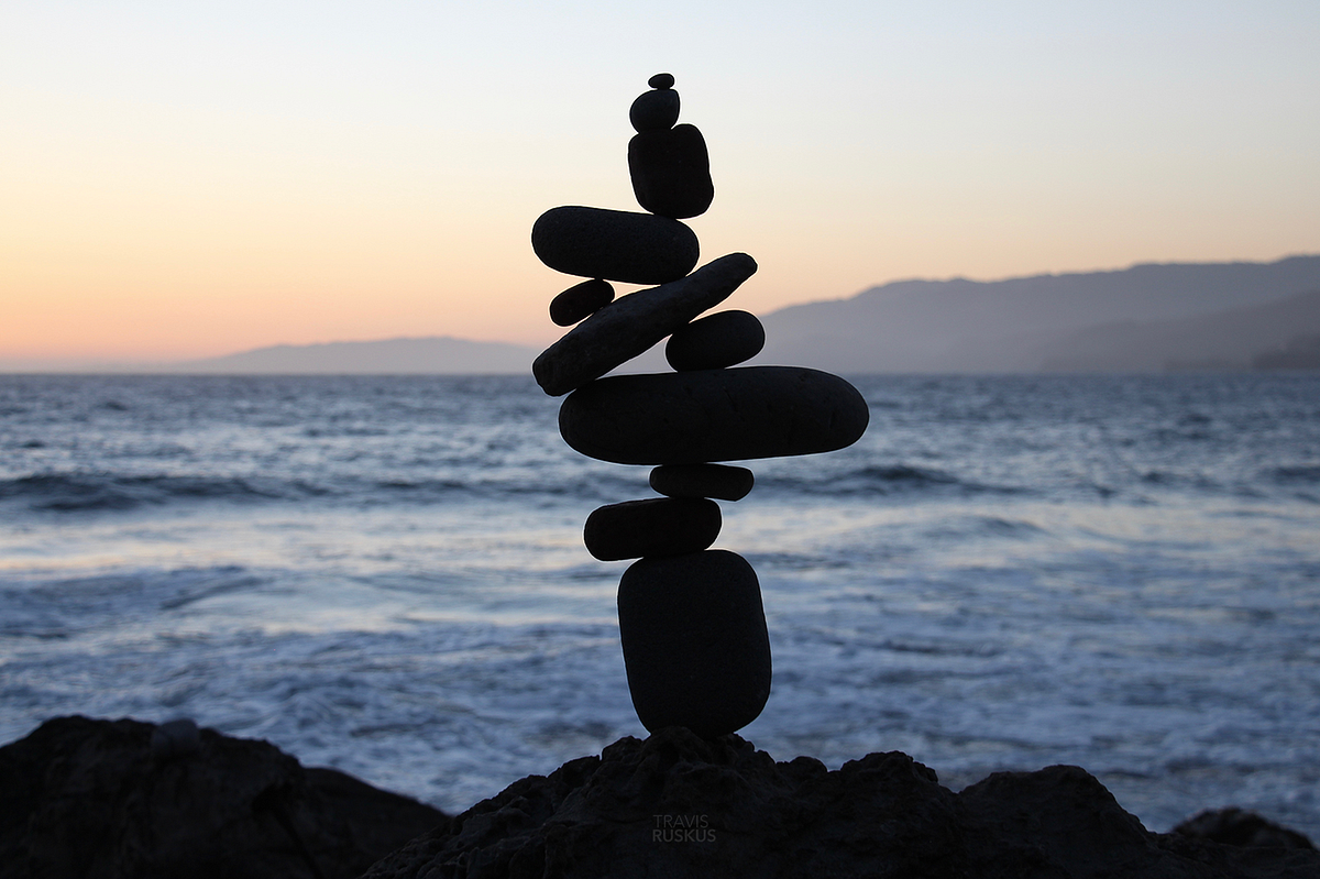 Una pila equilibrata di rocce lisce e scure si staglia contro l'oceano al tramonto, evocando la stabilità di portafogli resilienti 2026 mentre le onde morbide e le montagne lontane creano uno sfondo sereno.