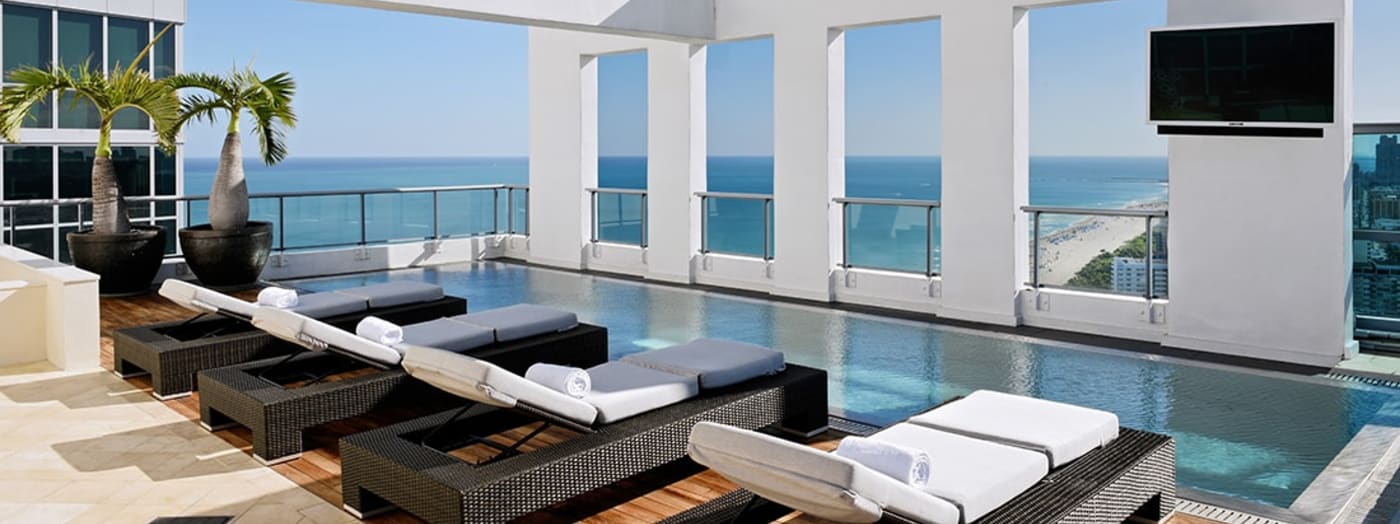 Modern rooftop pool area with lounge chairs, potted palm trees, large windows overlooking the ocean, glass railings, and a wall-mounted TV. The setting is sunny and luxurious with a clear blue sky.