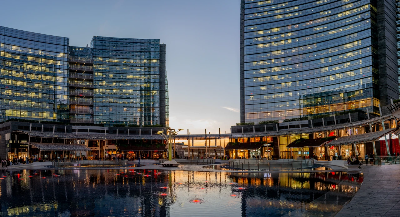 - mi prepari testo alternativo ,titolo , didascalia e descrizione per questa immagine ,? sempre per stesso articolo Grattacieli della finanza a Milano al tramonto, luci riflesse nell’acqua: Borsa in festa, listino che si assottiglia