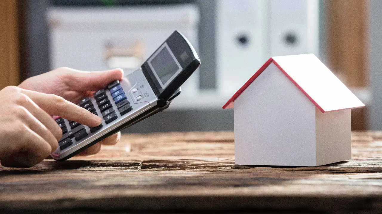 La mano di una persona utilizza una calcolatrice accanto a un modellino di casa su un tavolo di legno, suggerendo calcoli relativi alle finanze della casa o alle proprietà immobiliari.