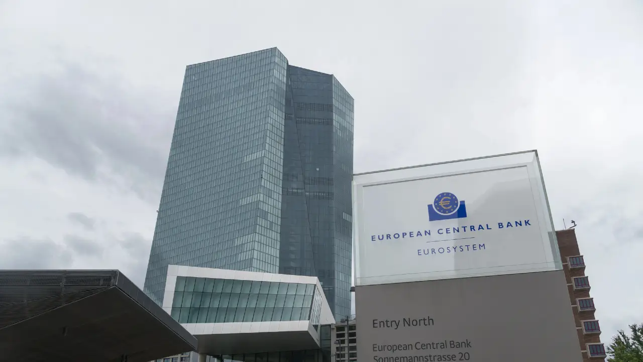 Un moderno grattacielo di vetro si erge dietro l'insegna della Banca Centrale Europea sotto un cielo nuvoloso. L'insegna mostra il logo della banca e recita "Banca centrale europea Eurosistema".