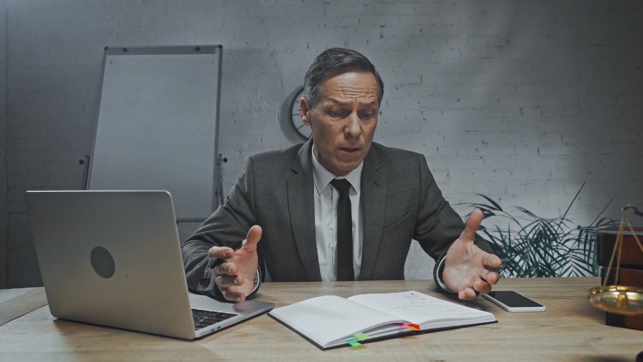 A man in a suit sits at a desk with a laptop, open notebook, and phone, gesturing with his hands in a concerned manner. A whiteboard and some plants are visible in the background.