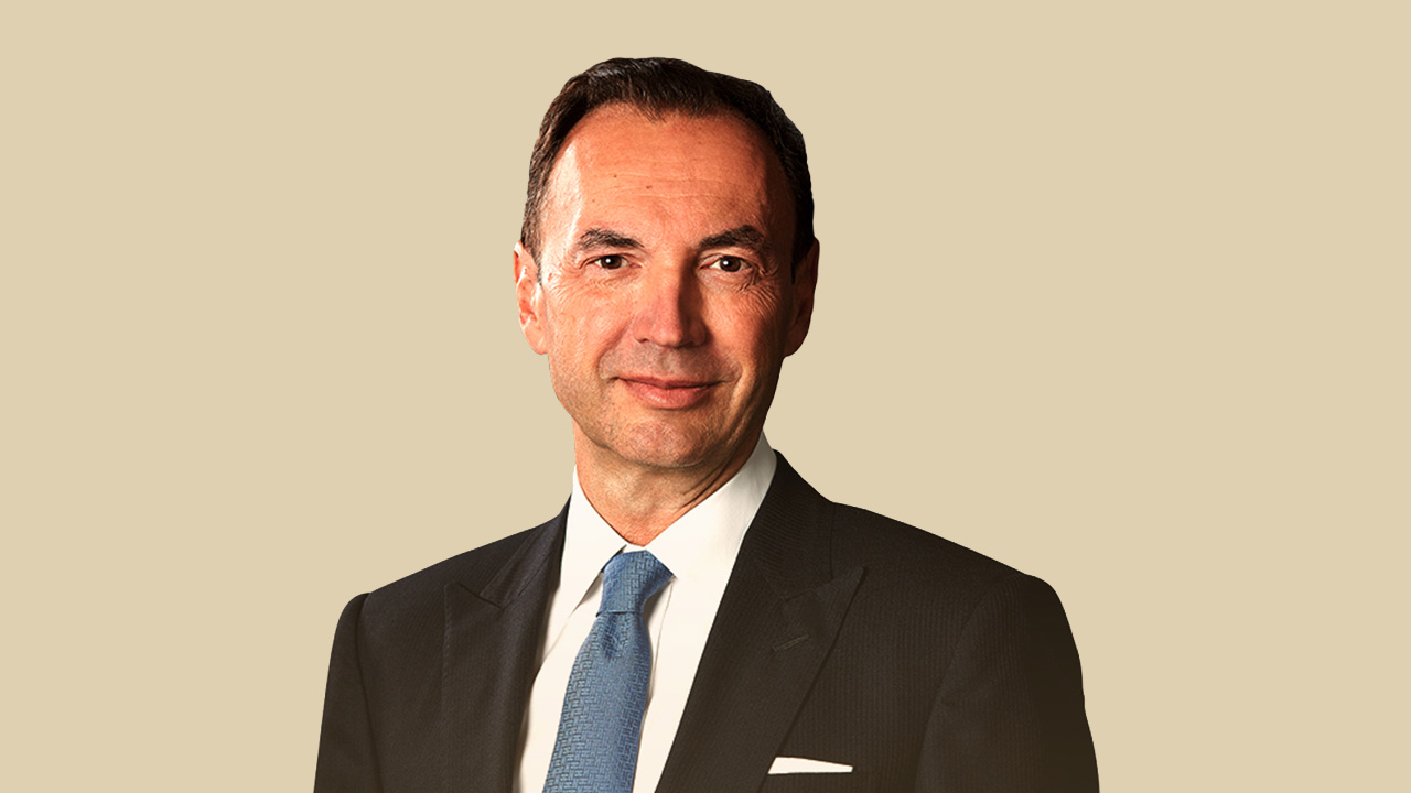 Un uomo in abito scuro e cravatta blu sorride dolcemente, su un semplice sfondo beige, incarnando la sicurezza delle scelte strategiche tipiche delle famiglie imprenditoriali.