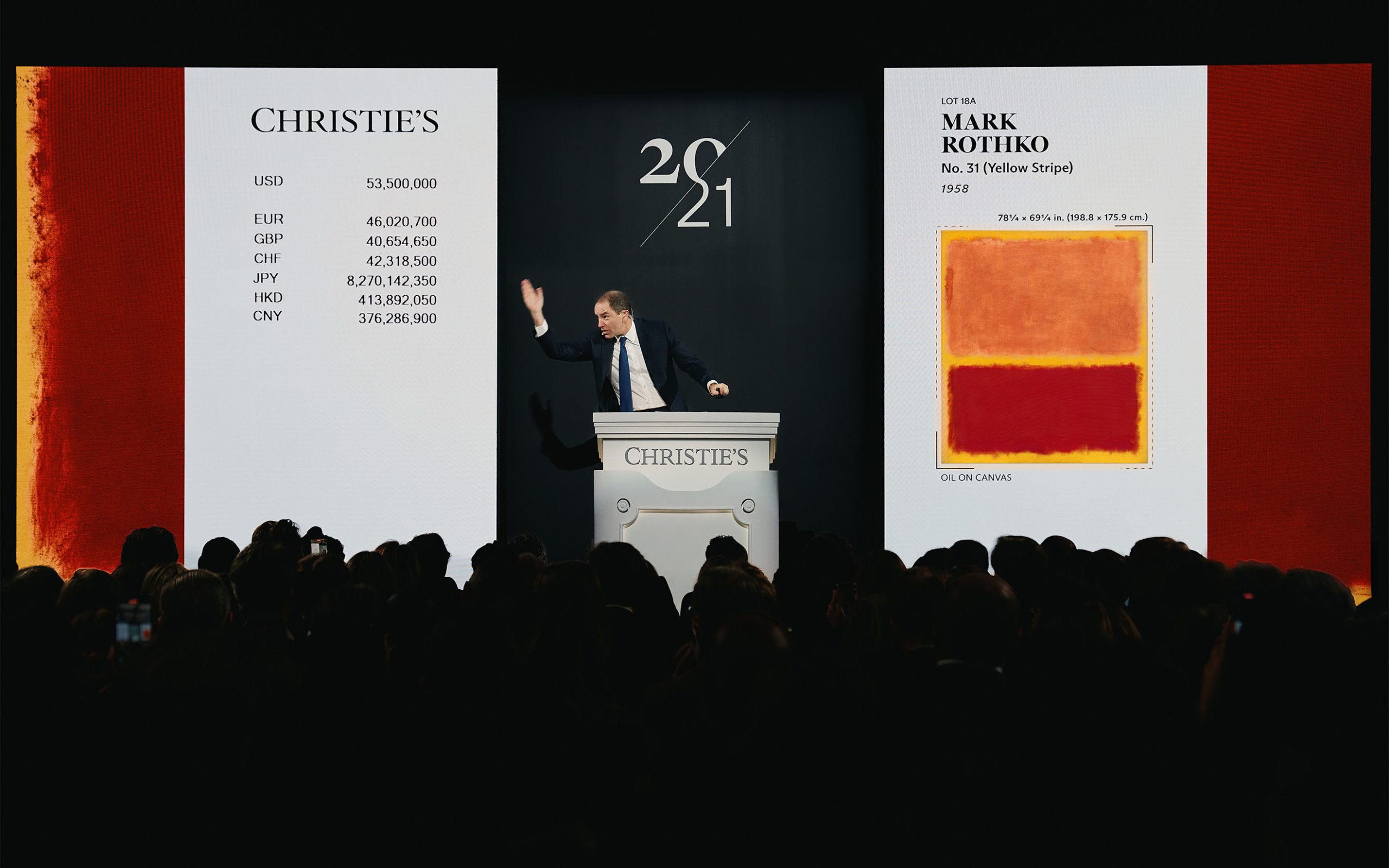 Un banditore d'asta si trova su un podio con un'immagine proiettata del dipinto di Mark Rothko "No. 7 (Yellow Stripe)" e il suo prezzo di vendita in varie valute durante un'asta d'arte di Christie's, di fronte a un pubblico seduto.