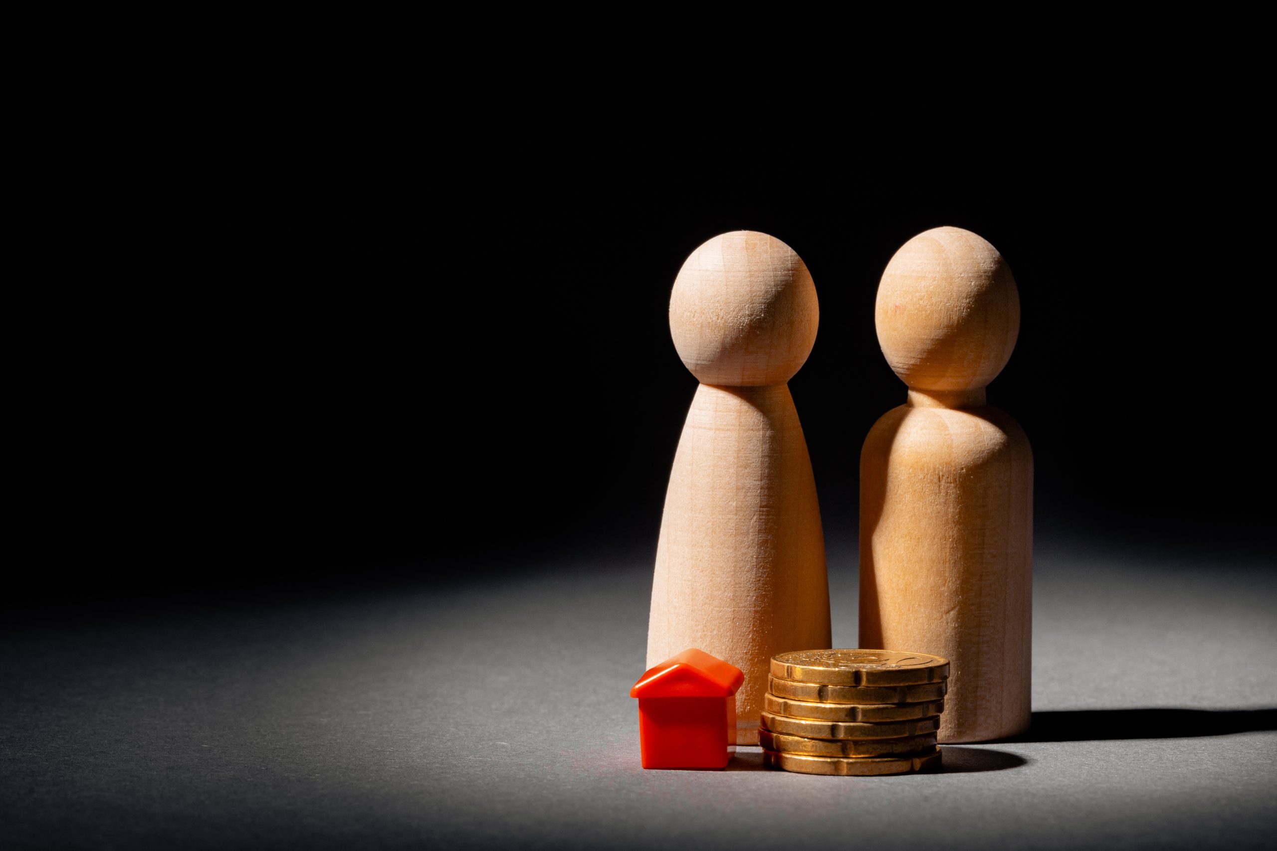 Due figure umane in legno si trovano accanto a una casetta giocattolo rossa e a una pila di monete, che simboleggiano concetti quali le finanze familiari, la casa, gli investimenti immobiliari o l'imposta successione Italia. La scena è ambientata su uno sfondo scuro.