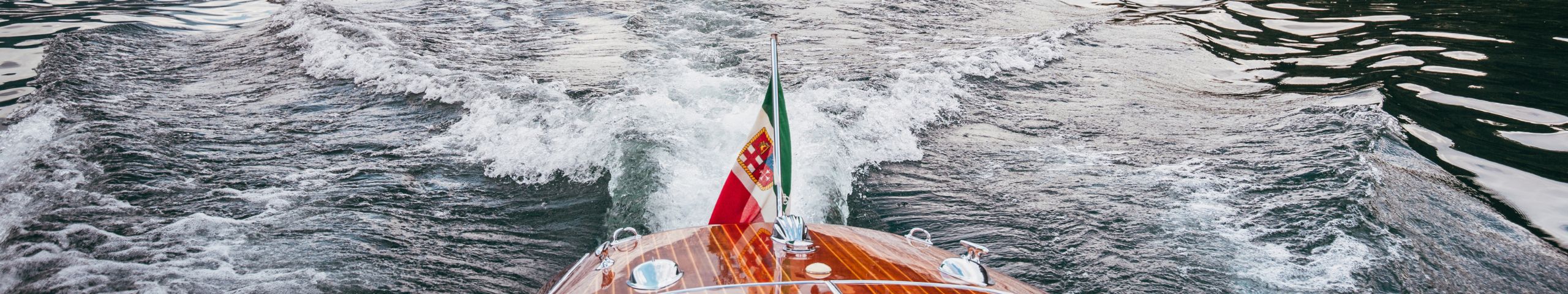 A wooden boat with a Portuguese flag sails on water, leaving foamy waves and ripples in its wake. The photo is taken from the boat’s stern, looking back at the water.