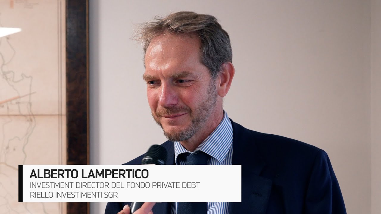 Un uomo in giacca e cravatta viene intervistato con un microfono. Sulla parete alle sue spalle è parzialmente visibile una mappa. Un testo in sovrimpressione in basso a sinistra recita: "Alberto Lampertico, Direttore Investimenti del Fondo Private Debt, Riello Investimenti SGR.