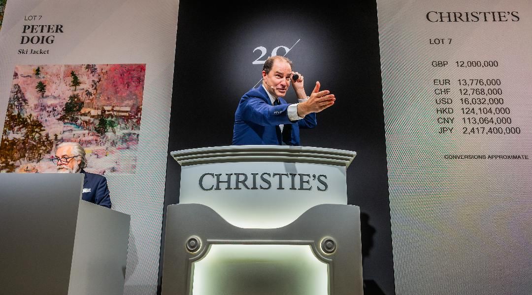 An auctioneer gestures while conducting a sale at a Christie’s auction. Behind him, a painting by Peter Doig and lot details, including bids in various currencies, are displayed on large boards. Another person sits on the left.