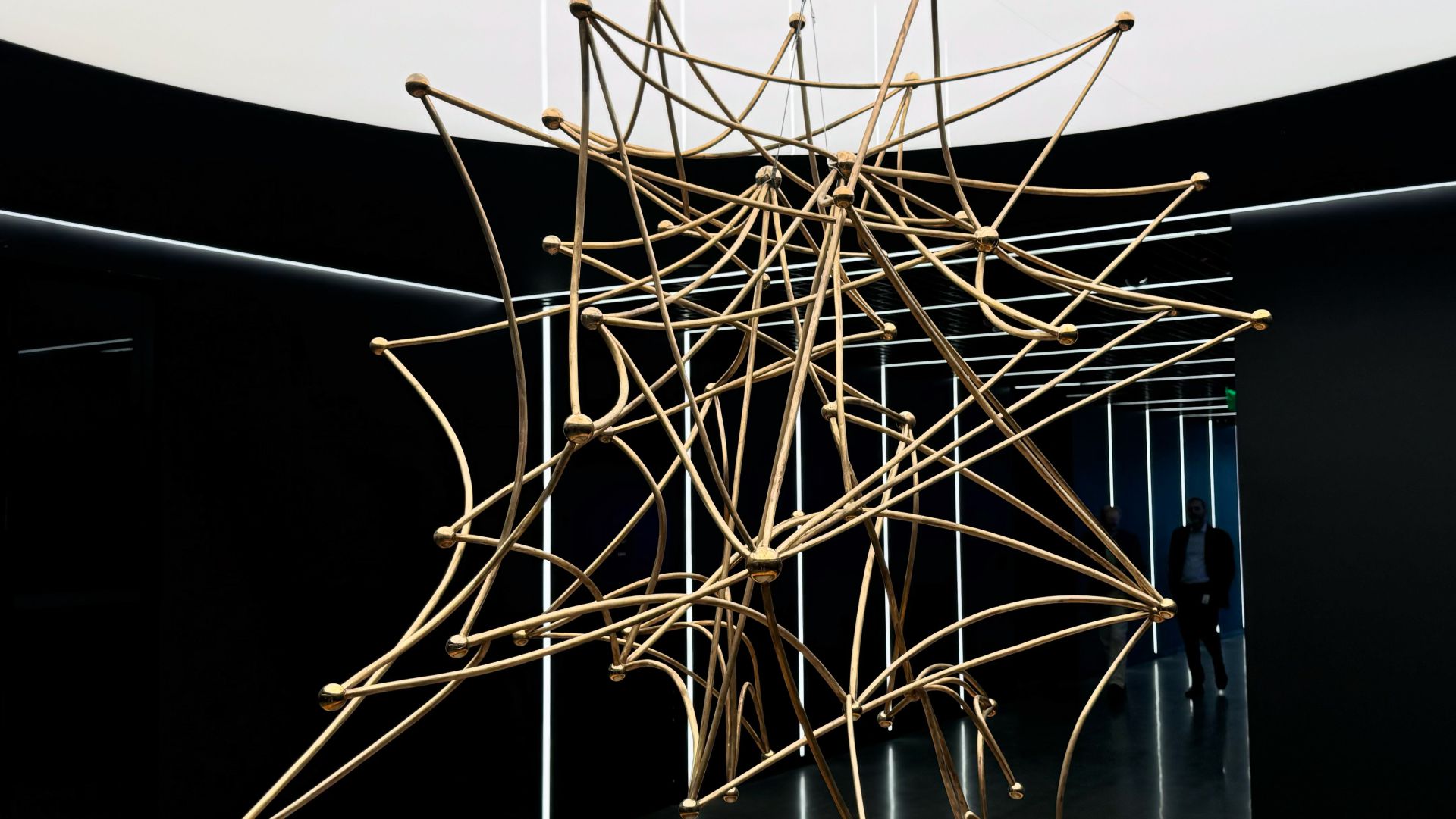 Una complessa scultura geometrica fatta di aste metalliche dorate che si intersecano è sospesa in un corridoio moderno con pareti scure e luci bianche sul soffitto. Sullo sfondo è visibile una persona.