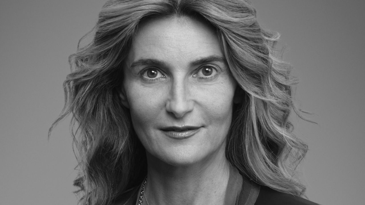Ritratto in bianco e nero di una donna con capelli ondulati lunghi fino alle spalle, che indossa un blazer e una collana, che sorride leggermente e guarda direttamente verso l'obiettivo su uno sfondo semplice.