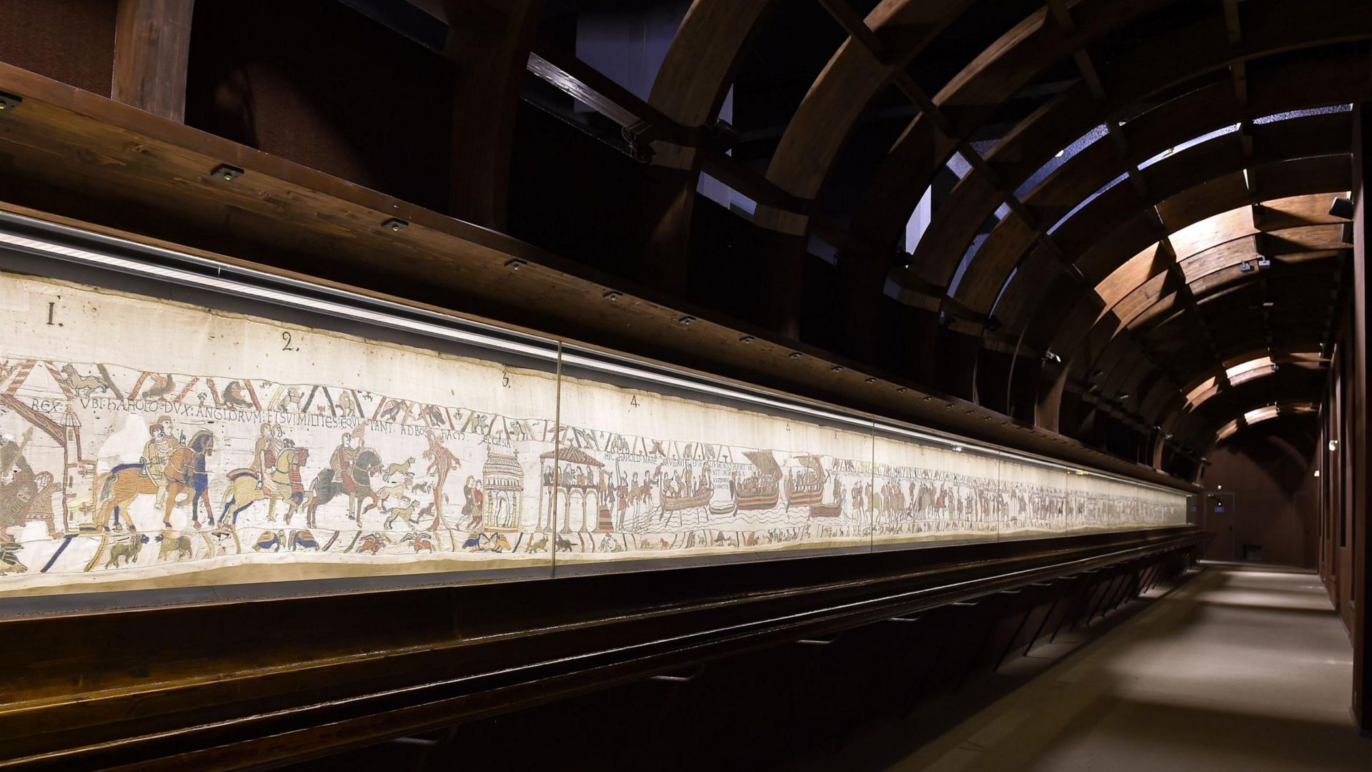 Una lunga esposizione illuminata dell'Arazzo di Bayeux, ospitata in una galleria ad arco poco illuminata con travi in legno e pareti scure, che mostra scene medievali ricamate su tessuto.