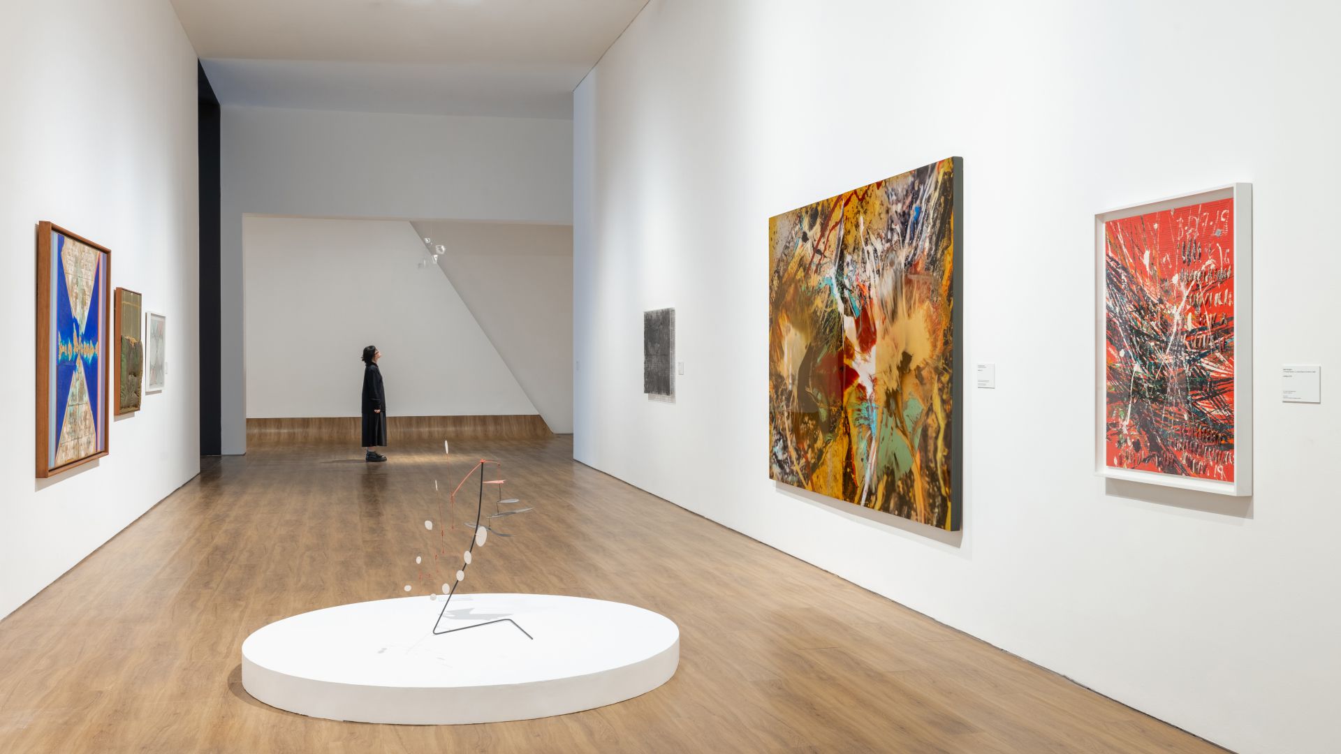 Una persona si trova in fondo a una spaziosa galleria d'arte con pavimenti in legno, pareti bianche, dipinti astratti e una moderna scultura mobile su una piattaforma circolare: un'atmosfera che ricorda le mostre del Max Mara Art Prize.