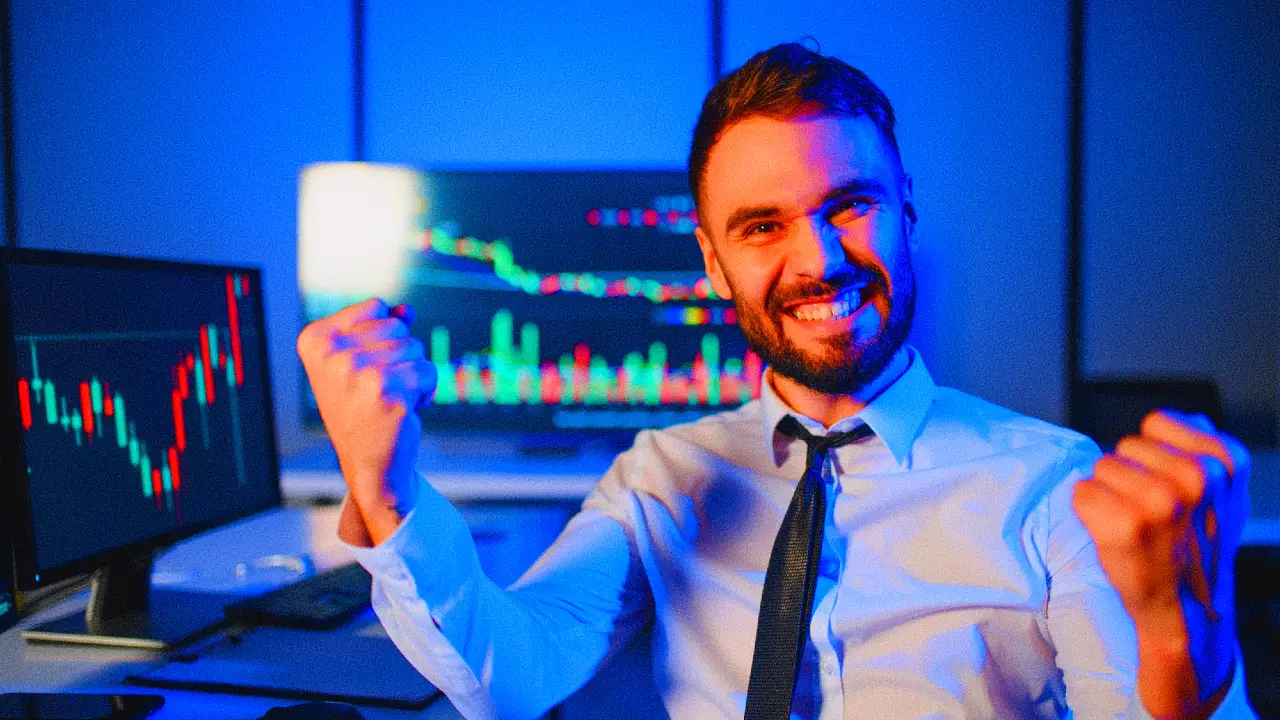 Un uomo sorridente in abiti professionali festeggia a pugni stretti davanti a monitor di computer che mostrano grafici colorati del mercato azionario. La scena è illuminata con luci blu.
