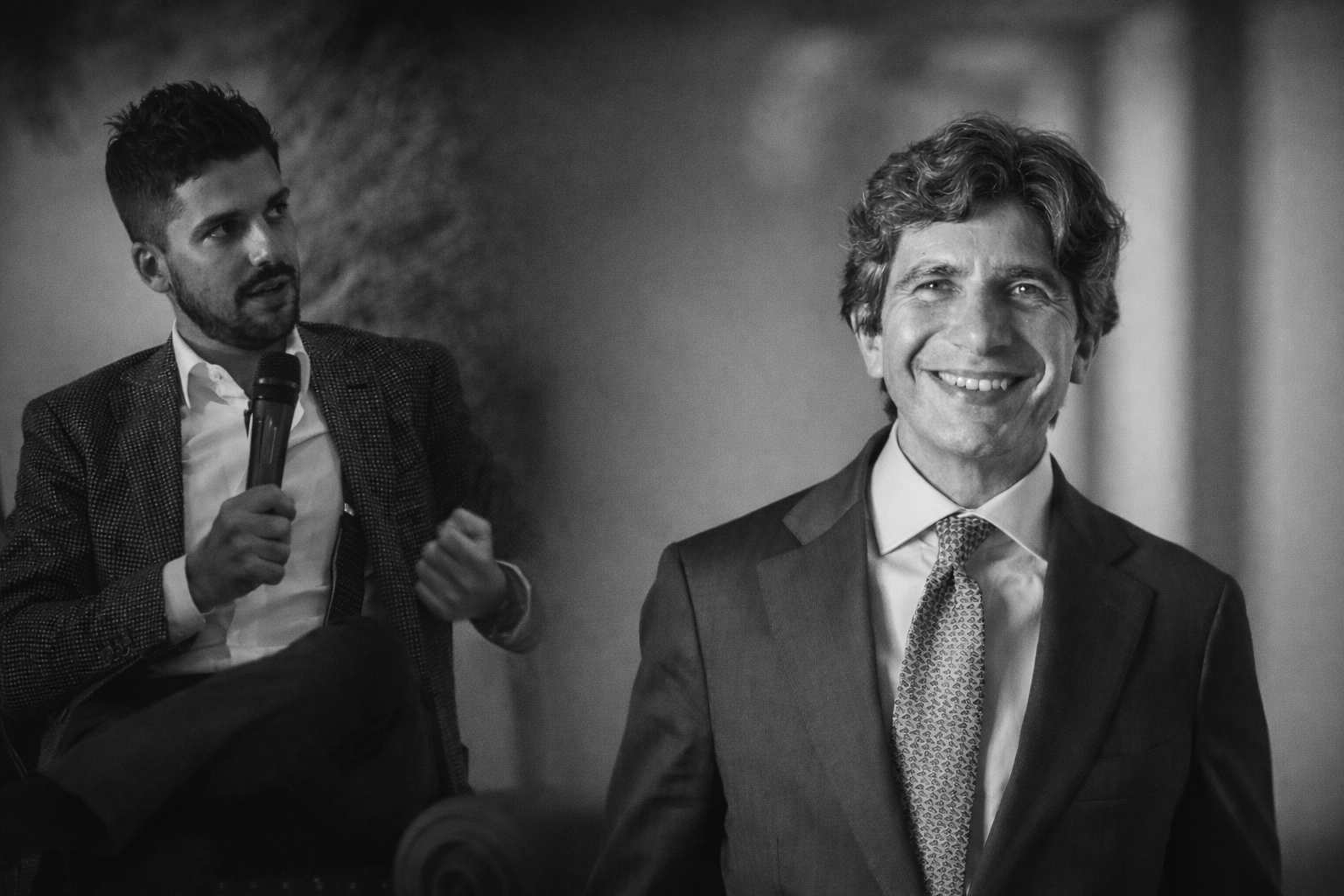Foto in bianco e nero di due uomini; uno è seduto e parla al microfono, indossando un blazer, mentre l'altro è in piedi in primo piano, sorridente e vestito in giacca e cravatta.