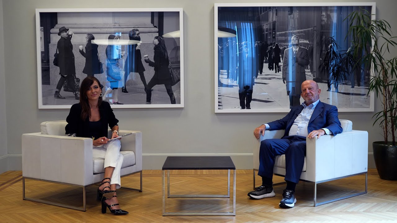 Paolo Colonna e Chiara Samorì siedono separate in poltrone bianche con un tavolino in mezzo, in una stanza moderna con pavimenti in legno e grandi foto in bianco e nero di scene di strada sulla parete alle loro spalle. Una pianta alta si trova in un angolo. Nuova puntata di Fondazioni in Dialogo