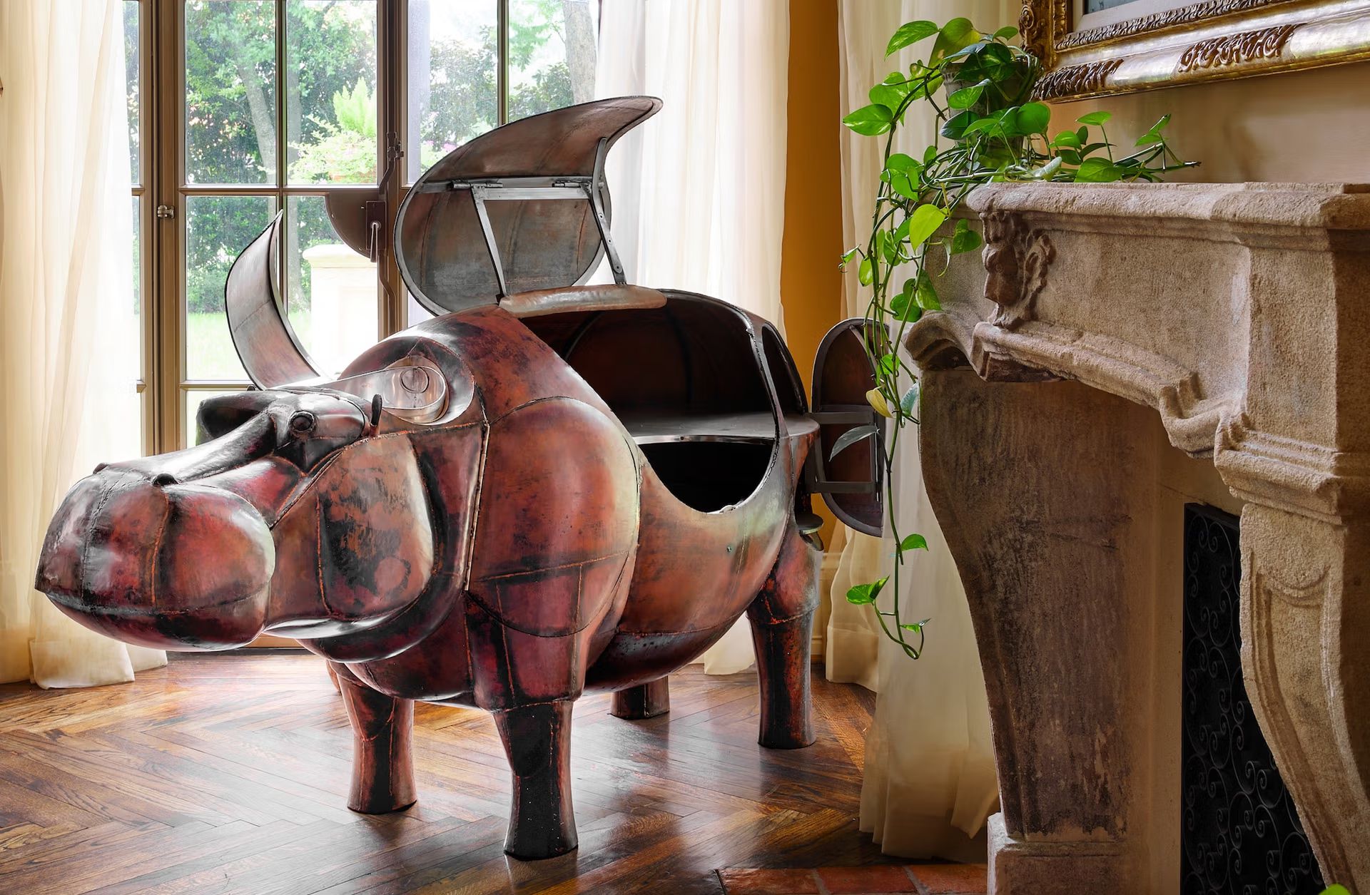 Una grande scultura metallica di un ippopotamo con scomparti aperti sul dorso e sul fianco si trova vicino a una finestra con tende velate, accanto a un camino in pietra e a una pianta verde e frondosa in una stanza accogliente.