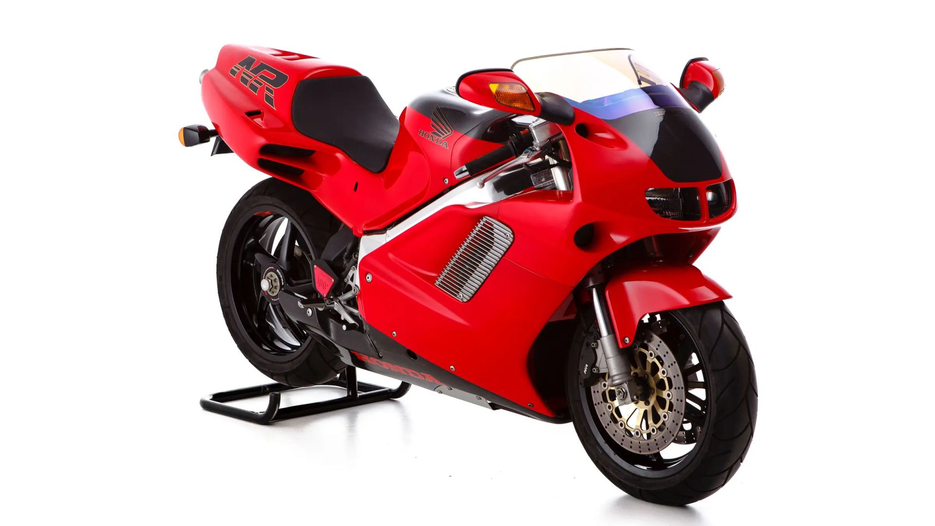 Una moderna moto sportiva di colore rosso brillante con accenti neri è esposta su un cavalletto posteriore su uno sfondo bianco. Il design è caratterizzato da carenature aerodinamiche, parabrezza trasparente e grandi freni a disco sulla ruota anteriore.