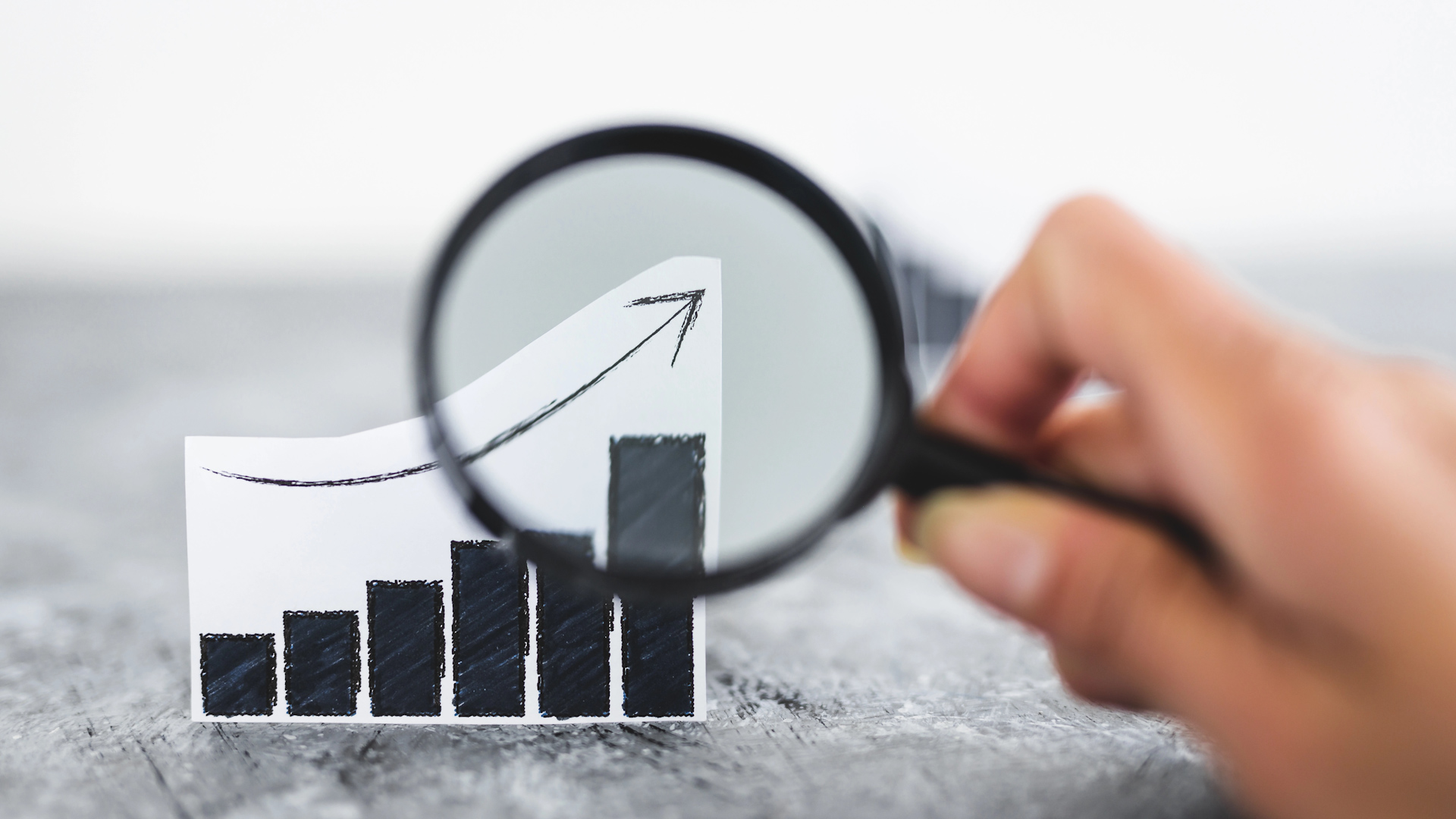 A hand holds a magnifying glass over a paper bar chart with an upward arrow, emphasizing growth and analysis. The background is blurred gray, focusing attention on the chart and magnifying glass.