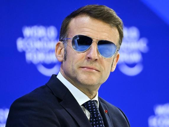 Un uomo in abito scuro, cravatta e occhiali da sole blu a specchio siede davanti a uno sfondo blu con testo bianco sfocato che recita "World Economic Forum". Guarda con sicurezza di lato.