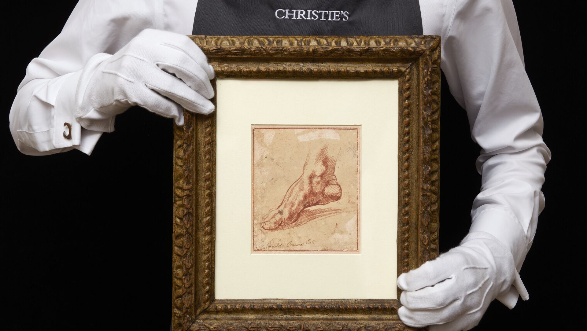 Una persona che indossa guanti bianchi e un grembiule da Christie's tiene in mano un disegno incorniciato di un piede umano, forse ispirato a Michelangelo Scoperto. L'opera d'arte è centrata da un'ampia stuoia color crema e da una cornice marrone ornata su sfondo nero.