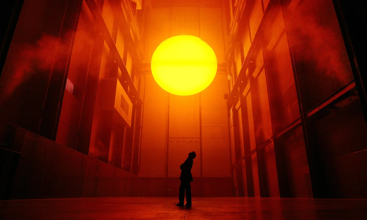 Una persona si trova in silhouette all'interno di un grande spazio industriale, con lo sguardo rivolto a una gigantesca sfera gialla e incandescente sospesa nell'aria, circondata da una luce arancione e nebulosa che riempie la stanza.