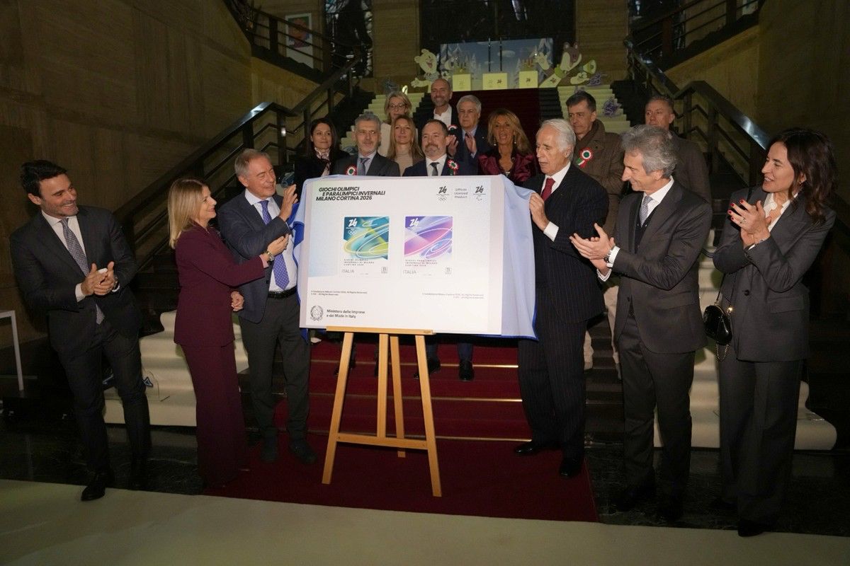 Un gruppo di persone in abiti formali si trova su una scalinata, riunito intorno a un grande tabellone con due francobolli commemorativi.