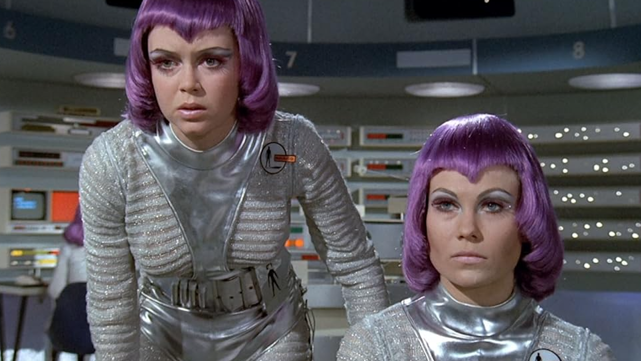 Due donne in uniformi futuristiche argentate e lucenti con parrucche viola siedono in una sala di controllo fantascientifica e retrò, piena di pulsanti e schermi. Una è in piedi e si sporge in avanti, l'altra è seduta, entrambe con un'espressione seria.