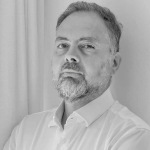 Un uomo con capelli corti e barba, che indossa una camicia bianca con colletto, è in piedi contro una parete e una tenda di colore chiaro e guarda direttamente la macchina fotografica con un'espressione neutra. La foto è in bianco e nero.