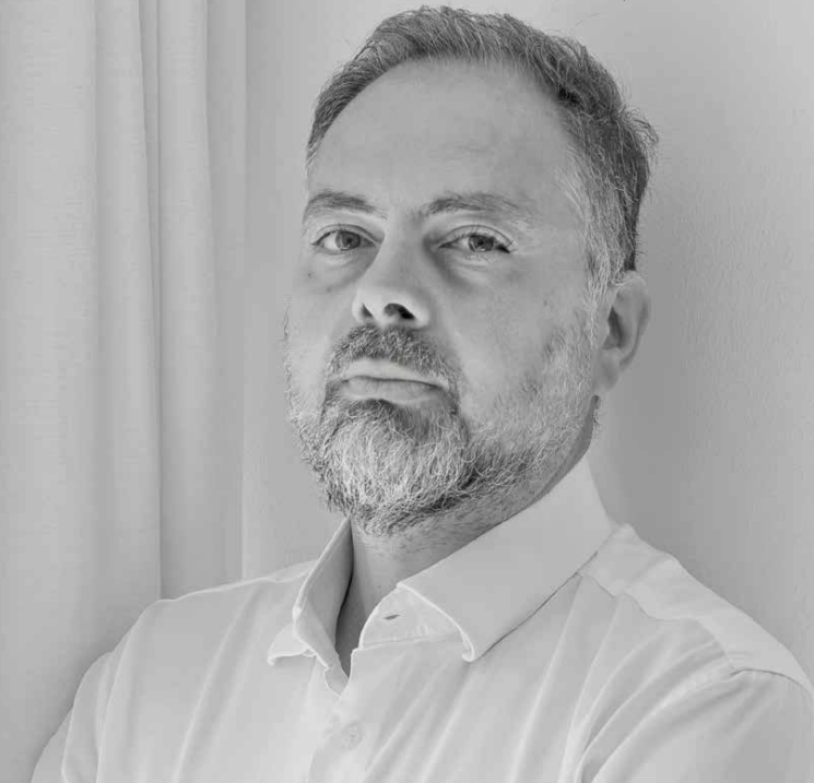 Un uomo con capelli corti e barba, che indossa una camicia bianca con colletto, è in piedi contro una parete e una tenda di colore chiaro e guarda direttamente la macchina fotografica con un'espressione neutra. La foto è in bianco e nero.