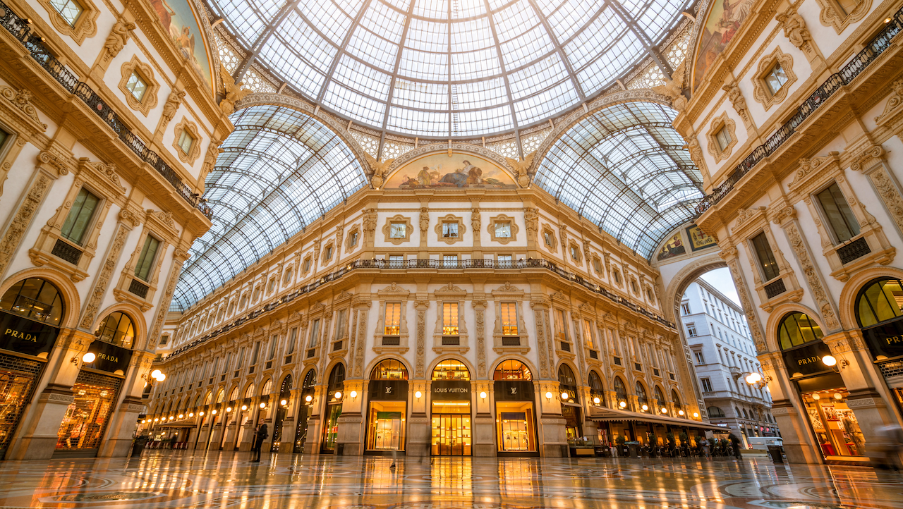 Elegante galleria commerciale al coperto a Lugano con architettura ornata, soffitto ad arco in vetro, pavimento in piastrelle decorative e illuminazione calda. Le vetrine e i negozi di lusso sono allineati alle pareti, sotto dettagli intricati e grandi finestre.