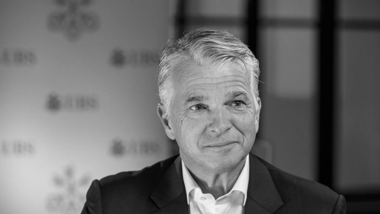 Un uomo con i capelli grigi che indossa un abito e una camicia bianca sorride leggermente guardando di lato; sullo sfondo sfocato il logo di UBS.