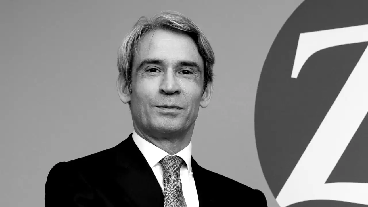 Un uomo in giacca e cravatta si trova davanti a un muro con una grande lettera "Z" parzialmente visibile sullo sfondo. L'immagine è in bianco e nero.